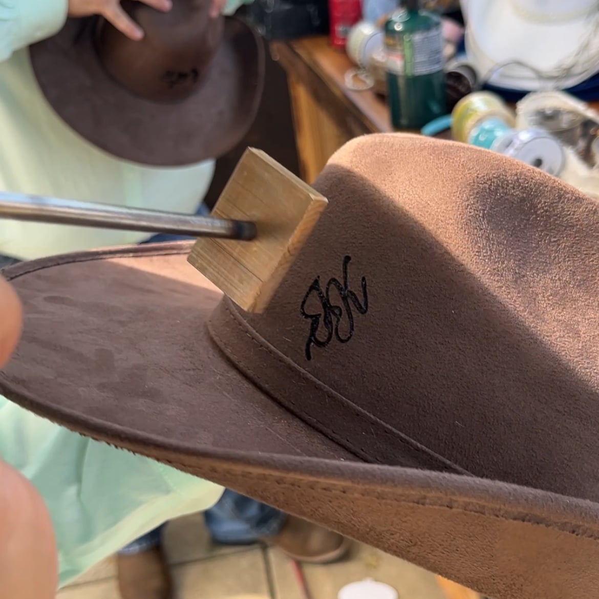 A branding iron on its wooden block resting on the crown of a tan felt hat, with a burned monogram on the side